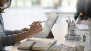 Side view of young professional designer concentrating on her task with digital tablet and painting tools on white desk in co working space