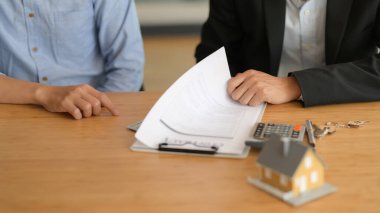 Emlak acentesinin, ev modeli ve ahşap masadaki malzemelerle müşteriye sözleşmeyi açıklarken çekilmiş görüntüsü.