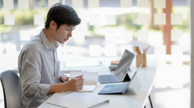Genç erkek girişimcilerin, cam duvar ofisindeki beyaz ahşap masa üzerinde dizüstü bilgisayar, tablet ve diğer ofis malzemelerini kullanırken işine odaklanmalarının yan görüntüsü 