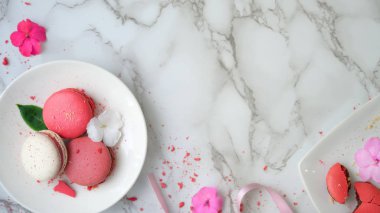 Fotokopi alanı beyaz seramik tabakta renkli pastel makarnaların üst görüntüsü, pembe kurdele ve mermer masa arkasında süslenmiş çiçek.