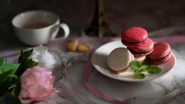 Close up view of Colourful Pastel Macarons on white ceramic plate with tea cup, pink ribbon and flower decorated on marble desk