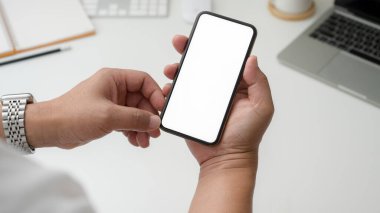 Beyaz masadaki boş ekranlı akıllı telefonu asgari çalışma alanındaki laptop ve ofis malzemeleriyle kesilen iş adamı görüntüsü.