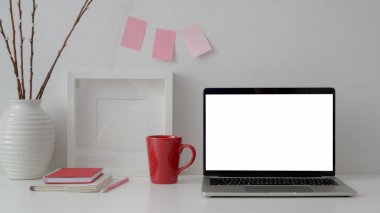 Boş ekran dizüstü bilgisayar, kitaplar, kırmızı bardak ve beyaz masa üzerindeki süslemeler ve beyaz duvardaki yapışkan notla şık işyeri görünümünü kapat 