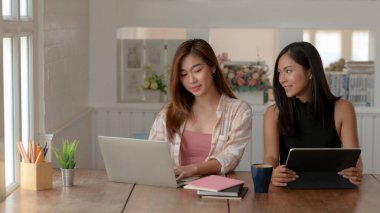 İki genç üniversiteli kızın modern oturma odasındaki dizüstü bilgisayar, tablet ve kırtasiye malzemeleriyle beyin fırtınası yapmalarına yakından bakın.