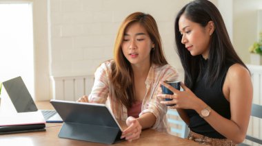 İki üniversiteli genç kızın modern oturma odasında tabletle birlikte kahve içerken çekilmiş fotoğrafları.