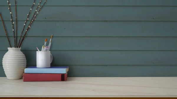 Close up view of simple artist workspace with painting brush, books, ceramic vase and copy space on marble desk with blue plank wall background