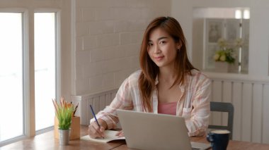 Modern oturma odasında dizüstü bilgisayar ve kırtasiyeyle ödevi üzerinde çalışan mutlu üniversiteli kızın kırpılmış görüntüsü. 