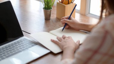 Üniversiteli genç bir kızın not defterine kırtasiye malzemesi ve ahşap masadaki dizüstü bilgisayarla kısa notlar almasına yakından bakın. 