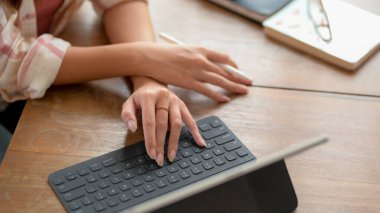 Üniversiteli genç bir kızın dijital tablette daktilo kullanmasını yakın çekim. Tahta masada, gözlüklü ve defterli. 