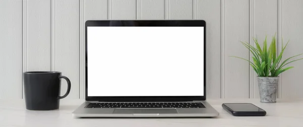 白い板の壁の背景と白いテーブルの上に空白の画面のラップトップ スマートフォン コーヒーカップと木の鍋で最小限のワークスペースの作物のショット — ストック写真