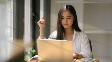 Genç üniversite öğrencisinin, kahve dükkanındaki ahşap masada dizüstü bilgisayarla ödevine odaklanmasını yakından izle.