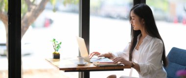 Bilgisayarında kırtasiye malzemesi ve kahve masasında kahve fincanı yazan genç üniversite öğrencisinin kırpılmış görüntüsü.