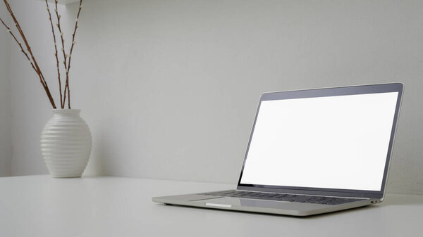 Cropped shot of minimal workspace with open blank screen laptop, vase and copy space on white table with white wall 