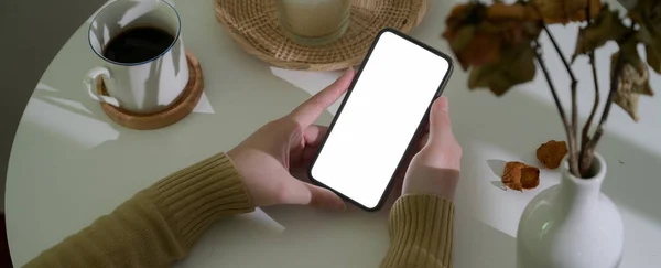 Close up view of a woman sitting at workspace and using blank screen smartphone with coffee cup and decorations on white circle table