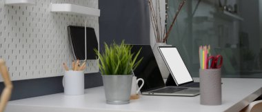 Cam bölme odasındaki beyaz masa üzerinde sahte dijital cihazlar, malzemeler ve dekorasyonlarla minimal ofis masasının yan görüntüsü 