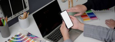 İki tasarımcının projelerini tartışırken beyaz masa üzerinde akıllı telefon, dizüstü bilgisayar ve tasarımcı malzemeleriyle çekilmiş görüntüler. 