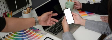 Tasarımcı ekibinin projelerini beyaz masa üzerinde akıllı telefon, dizüstü bilgisayar ve tasarımcı malzemeleriyle tartışırken çekilmiş görüntüleri. 