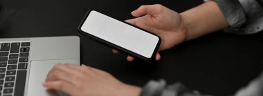 Karanlık, modern ofis masasında çalışan akıllı telefon ve dizüstü bilgisayarlı kadın girişimcinin yan görüntüsü.