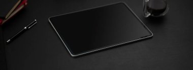 Siyah masa üzerinde dijital tablet, kahve fincanı ve diğer malzemelerle dolu koyu modern çalışma alanı.  