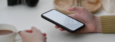 Kadın serbest çalışanın, müşteriyle iletişime geçmek için akıllı telefon kullanmasını ve beyaz masada sıcak kahve içmesini yakından izleyin.