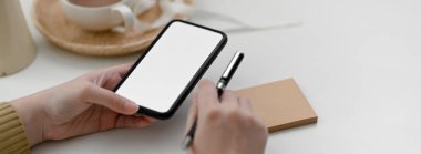 Kadın serbest çalışanın, kahve fincanı ve not defteriyle beyaz masadaki müşteriyle bağlantı kurmak için akıllı telefon kullanmasını kapat.