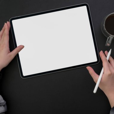 Boş ekran tableti üzerinde çalışan serbest çalışan, koyu modern çalışma alanında stilus kalem ve kahve fincanı kullanan kadın.