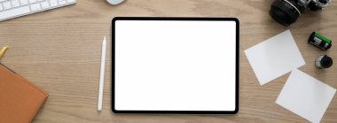 Boş ekran tableti, kamera, not defteri ve ahşap masadaki malzemelerle rahat çalışma alanının üst görüntüsü  