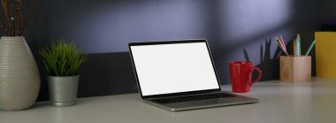 Boş ekran dizüstü bilgisayarı, kırtasiyesi ve gri tavan arası duvarlı beyaz masa dekorasyonu olan şık bir ev ofisinin kırpılmış görüntüsü.