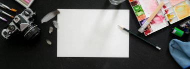 Eskiz kağıdı, boyama araçları, kamera ve diğer malzemeler ile sanatçının çalışma alanının üst görünümü siyah masa üzerinde 
