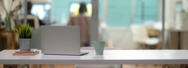 Dizüstü bilgisayarı, kupası, ağaç kabı, ofis malzemeleri ve cam bölme ofisindeki beyaz masa üzerindeki fotokopi alanı ile rahat ofis masasını kapatın.