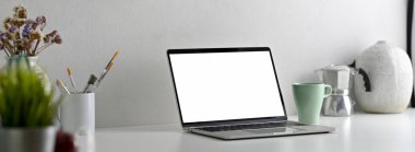 Boş ekran dizüstü bilgisayarlı modern ofis masasının yan görünümü, beyaz masadaki resim araçları ve dekorasyonlar