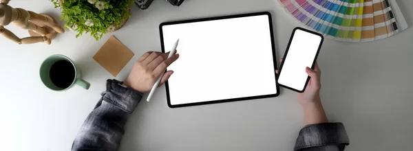 Top view of female freelancer working on her project with mock-up tablet, smartphone and designer supplies on white table