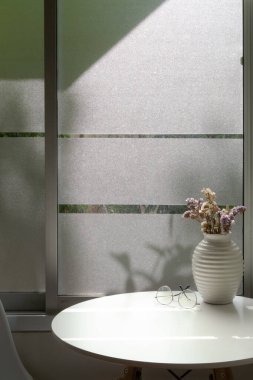 Pencerenin yanındaki kahve sehpasını, vazo, gözlük ve beyaz yuvarlak masa üzerindeki fotokopi alanını kapat.   