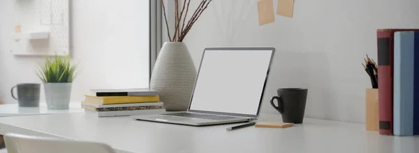 Cropped shot of portable workspace with mock-up laptop, mug, books, notepad and decoration on white desk