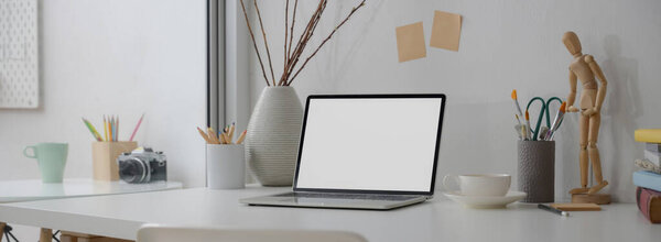 Cropped shot of designer workspace with mock-up laptop, designer supplies, cup and decoration on white desk 