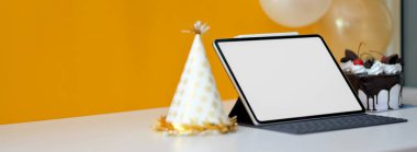 Çevrimiçi serebrasyon kavramı, boş ekran tableti ve parti şapkası beyaz masada, dekorasyonlar sarı arka planda.