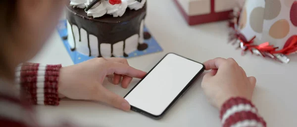 Online Birthday celebration concept, female using blank screen smartphone on white table with cake, present box and decorations