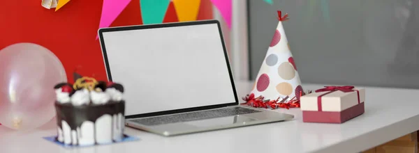 Online Birthday celebration concept with blank screen laptop, cake and decorations on white table with red wall