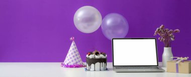 Çevrimiçi doğum günü partisi konsepti boş ekran dizüstü bilgisayar, pasta, parti şapkası ve mor duvarlı beyaz masa süsleri