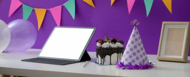 Çevrimiçi doğum günü partisi konsepti boş ekran tableti, pasta ve parti şapkası beyaz masada mor duvarda süslemelerle