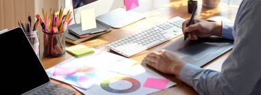 Erkek grafik tasarımcısının, ahşap ofis masasında tablet, bilgisayar ve evrak çizerken çekilmiş görüntüleri. 