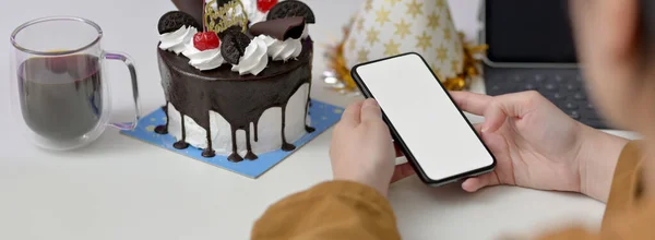 Online birthday party concept, female using mock-up smartphone on white table with cake and party hat