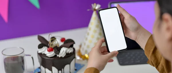 Online birthday party concept, female video calling on mock-up smartphone at white table with cake and party hat