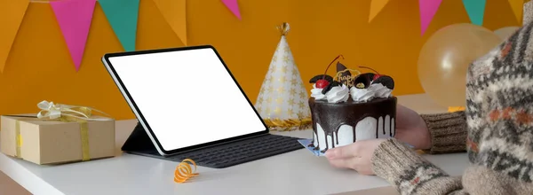 Online birthday party concept with blank screen tablet, cake, party hat and present box on white table with decorations on yellow wall