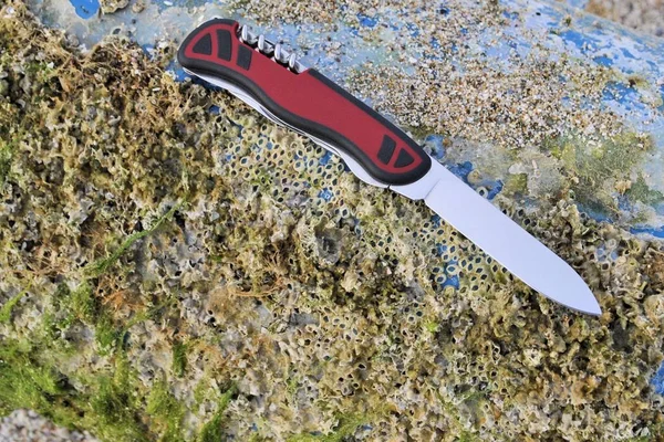 Multi function folding knife stainless blade black red handle travel vacation beach stone background