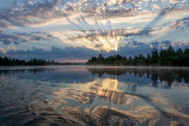 Nehirde gün batımı. Güneş ışınları bulutları delip geçiyor. Sudaki yansıma..