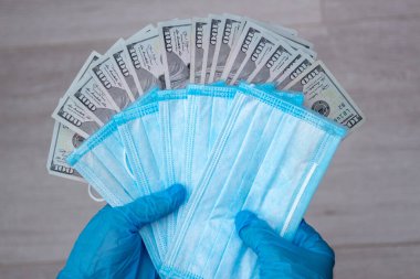 Tıbbi lateks eldiven giyen ellerde yüz dolarlık banknotlar ve birkaç mavi tek kullanımlık tıbbi maske var. Yakın plan, üst görüş. İlaç maliyeti kavramı