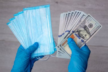 Tıbbi lateks eldiven giyen ellerde yüz dolarlık banknotlar ve birkaç mavi tek kullanımlık tıbbi maske var. Yakın plan, üst görüş. İlaç maliyeti kavramı