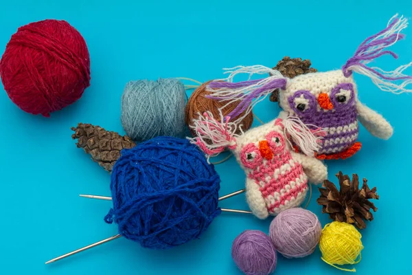 Knitted handmade small colored toys, colorful owls next to skeins of thread for needlework, knitting needles and dry pine cones on a blue background. Women\'s hobby and festive decor concept