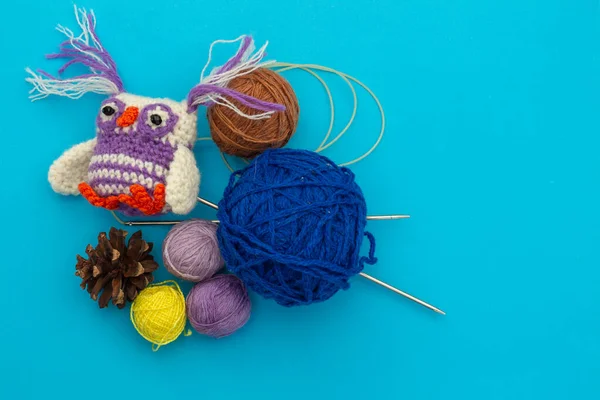 Knitted handmade small colored toy, colorful owlet next to skeins of thread for needlework, knitting needles and dry pine cones on a blue background.  Needlework and children\'s creativity concept